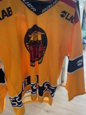 Gul Luleå Hockey matchtröja Bauer - Säljer en gul Luleå Hockey matchtröja från Bauer med klubbens logga framtill och flera sponsortryck. Tröjan har långa ärmar, svart och röd detalj upptill samt vita och svarta ränder nedtill. Perfekt för hockeyfans som vill stötta sitt lag.