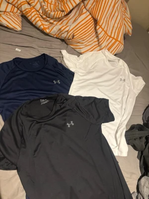  Armour tränings-tshirts mörk blå äkta - Säljer tre snygga tränings-tshirts från Under Armour i färgerna svart, marinblå och vit. Alla har rund hals och är tillverkade i ett lätt och snabbtorkande funktionsmaterial, perfekt för träning och sport.