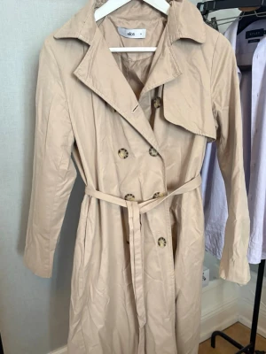 Beige trenchcoat från Ellos - Stilren beige trenchcoat från Ellos med klassisk dubbelknäppning och bälte i midjan. Jackan har bred krage, långa ärmar och dekorativa knappar i sköldpaddsmönster. Perfekt för dig som vill ha en tidlös och trendig look.