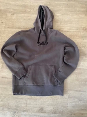 Grå hoodie från Hollister - Snygg grå hoodie från Hollister med svart broderad logga på bröstet och klassisk känguruficka. Mjuk insida och justerbar huva med svarta snören. Perfekt för en avslappnad och trendig stil. Kom med frågor eller prisförslag🤝