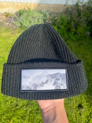 Svart ribbstickad mössa med patch från Revenite - Säljer en svart ribbstickad mössa med uppvikt kant och en stor patch framtill som visar ett svartvitt bergsmotiv med en person. Mössan har en clean och stilren look som passar till det mesta. Perfekt för dig som gillar streetwear och vill ha något unikt. Limiterad i 500