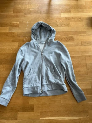 Grå zip hoodie från Moncler - Snygg grå hoodie från Moncler med dragkedja framtill och två fickor. Klassisk huva och ribbade muddar vid ärmslut och nederkant. Moncler-logga på ärmen ger en exklusiv touch. Perfekt till chill eller streetwear-stil. Finns lite problem att stänga den men funkar fortfarande 