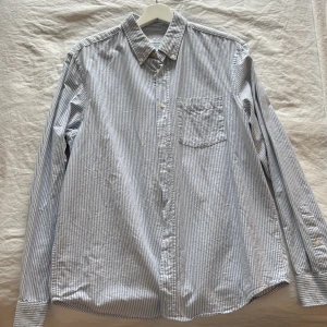 Randig skjorta från Zara - Klassisk blå- och vitrandig skjorta från Zara med bröstficka och knappar framtill. Skjortan har lång ärm och en avslappnad passform, perfekt till jeans eller chinos. Tillverkad i bomull för en skön känsla.