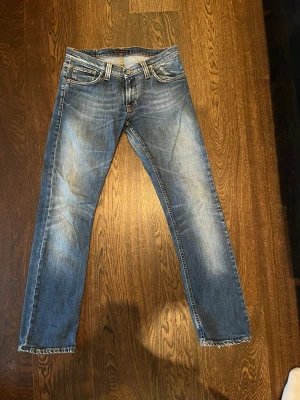 Blå raka jeans slitna från Nudie Jeans - Säljer ett par klassiska blå jeans från Nudie Jeans med raka ben och slitningar. En snygg tvättad look. Perfekta för en avslappnad stil.                Midja 87,5 cm när jeansen är knäppta.  Ben längd från grenen och ner 81 cm Ben längd från midjan och ner 104,5 cm.