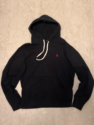 Svart hoodie från Polo Ralph Lauren - Klassisk svart hoodie från Polo Ralph Lauren med vit snören och röd logga på bröstet. Hoodien är tillverkad i bomulls material och är en perfekt hoodie som aldrig går fel att ha på sig. Kontakt mig gärna om du har fler frågor. Hoodien skickas ut samma dag eller dagen efter som den har beställts📦