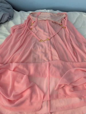 Rosa transparent klänning Ida Sjöstedt - Supersöt rosa klänning från Ida Sjöstedt i transparent, flytande material. Klänningen har en lös passform, dekorativa gulddetaljer vid halsen och ett tunt tyg som ger en drömmig look. Perfekt för dig som gillar romantisk och unik stil.