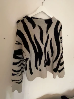 Stickad tröja med zebramönster - Säljer en stickad tröja med zebramönster i svart och beige. Tröjan har en lös passform, rund halsringning och långa ärmar. Perfekt för dig som gillar djurmönstrat och vill ha en mysig, trendig look.