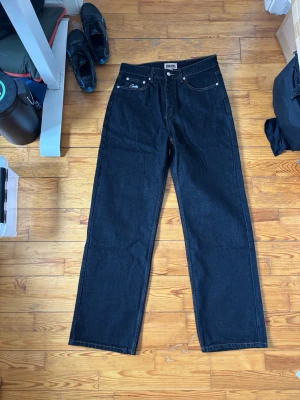 Svarta jeans från cortiez - Säljer ett par svarta raka jeans från cortiez med klassisk femficksdesign. Jeansen har en cool detalj med röd, vit och grön rand samt Cortiez-logga på ena bakfickan. Tillverkade i slitstark denim, perfekta för en streetwear-look.