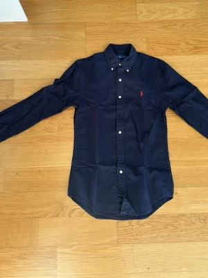 Marinblå skjorta från Ralph Lauren - Klassisk marinblå skjorta i slim fit från Ralph Lauren med button-down krage och långa ärmar. Skjortan har vita knappar framtill och den ikoniska röda logotypen broderad på bröstet. Tillverkad i mjuk bomull för en bekväm känsla. Använd ett fåtal gånger.