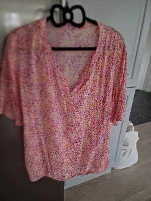 Blommig v-ringad blus i rosa och orange från Best of Basic - Säljer en luftig blus med v-ringning och korta ärmar. Blusen har ett färgglatt blommigt mönster i rosa, orange och vitt. Perfekt för varmare dagar och ger en fräsch look till din outfit. Köpte från Best of Basic. 