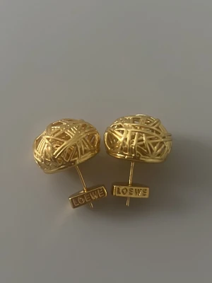 Örhängen från Loewe - Unika örhängen från Loewe i guldfärgad metall med en cool, trådinspirerad design som påminner om små bollar av guldtråd. Märkets logga syns tydligt på plattan vid stiftet. Perfekta för dig som gillar statement-smycken med modern känsla.