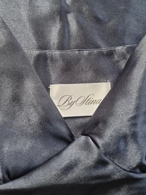 Marinblå klänning  i satin By Stina - Snygg marinblått klänning/balklänning från By Stina i glansigt satinmaterial. Klänningen har smala axelband och v-ringad urringning för en elegant och bekväm känsla. Perfekt för avslutningsfester eller baler. Inköpt 2025. Använd endast en kväll. 