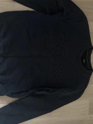 Svart sweatshirt från BOSS - Snygg mörkblå sweatshirt från BOSS med stor ton-i-ton logga broderad på bröstet. Tröjan har rund halsringning och långa ärmar.