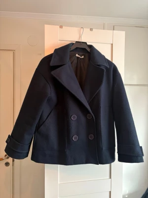 Mörkblå dubbelknäppt ullrock - Kort mörkblå rock med dubbelknäppning och bred krage. Rocken har två rader stora knappar framtill och dekorativa knappar vid ärmsluten. Tillverkad i ett tjockare ullmaterial och helfodrad för extra värme.