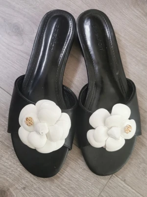 Svarta sandaler med vit blomma storlek 38 - Säljer ett par svarta sandaler från Joules med en stor vit blomapplikation framtill och guldfärgad detalj i mitten av blomman. Sandalerna har öppen tå och låg klack, tillverkade i konstläder med en stilren och trendig look.