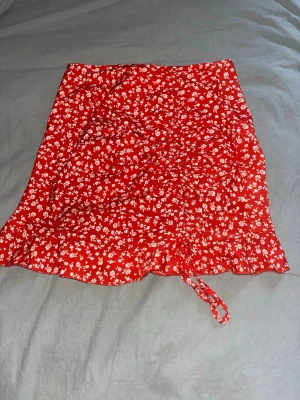 Röd blommig kortkjol med snörning - Söt röd kortkjol med vitt blommönster och snörning framtill. Kjolen har volangkant nedtill och är perfekt för varma dagar.