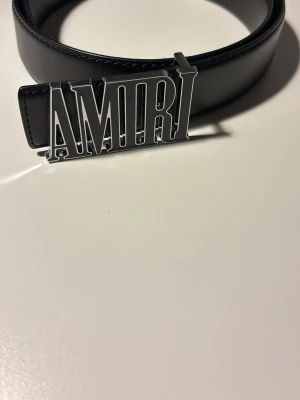 Svart skärp från Amiri med silverdetalj - Snyggt svart skärp från Amiri med en stor, iögonfallande silverspänne där det står AMIRI. Skärpet är gjort i slätt skinn och har en clean, modern look som passar till många olika outfits.