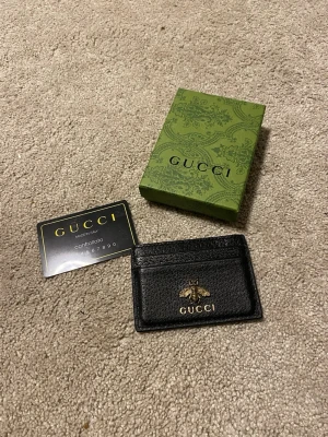 Svart läder korthållare med Bi - Snygg svart korthållare i äkta läder med guldfärgad bi-detalj och logotyp framtill. Kommer med box och kort. Perfekt för dig som vill ha stilren och lyxig accessoar till dina kort.