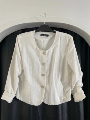 Vit linnéjacka från Mac Scott - Stilren vit jacka från Mac Scott i linne med rund halsringning och stora knappar framtill. Jackan har lång ärm och en lätt figursydd passform som ger en klassisk look. Perfekt för dig som gillar minimalistisk stil. 80-talet