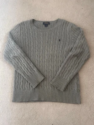 Grå kabelstickad tröja Polo Ralph Lauren - Kabelstickad grå tröja från Polo Ralph Lauren med rund halsringning och diskret broderad logga på bröstet. Tröjan har långa ärmar och breda ribbade muddar vid ärmslut och nederkant. Perfekt för dig som gillar klassisk och stilren look. Tveka inte vid att ställa frågor.
