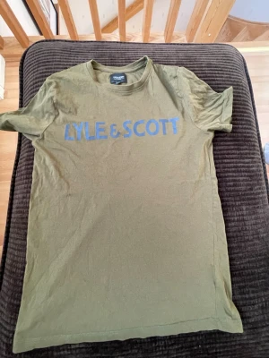 Olivgrön Lyle & Scott t-shirt - Snygg olivgrön t-shirt från Lyle & Scott Junior med stort blått tryck på bröstet. Klassisk rund halsringning och korta ärmar. Perfekt för dig som gillar enkel och stilren design. Tillverkad i mjuk bomull för skön känsla hela dagen.