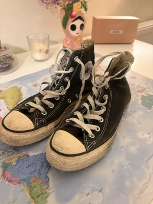 Svarta Converse All Star High - Klassiska svarta Converse All Star high-top sneakers med vit gummisula och tå, vita snören och den ikoniska loggan på sidan. Skorna är i canvasmaterial och har kontrastsömmar som ger en retrovibe. Perfekta för dig som gillar streetstyle och tidlös design.