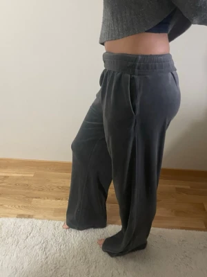 Mörkgrå velour mjukisbyxor - Säljer ett par mörkgrå mjukisbyxor i mjuk velour med bred resår i midjan och raka, lösa ben. Perfekta för chill dagar hemma eller när du vill vara extra bekväm. Byxorna har fickor på sidorna och en avslappnad passform.