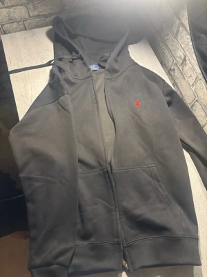 Svart hoodie från Polo Ralph Lauren - Säljer en svart hoodie från Polo Ralph Lauren med dragkedja och huva. Klassisk röd logga broderad på bröstet och stora fickor framtill. Tillverkad i mjukt bomullsmaterial och har ribbade muddar vid ärmslut och nederkant.