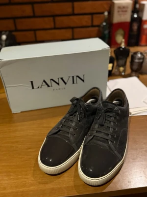 Svarta Lanvin sneakers i mocka och lack - Snygga svarta sneakers från Lanvin med ovandel i mocka och tåparti i blankt lack. Klassisk låg modell med vita sulor och svarta skosnören. Perfekta för dig som gillar stilrena och exklusiva sneakers med en twist.