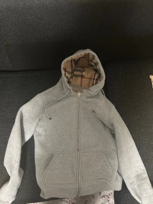 Grå hoodie från Burberry med dragkedja - Snygg grå hoodie från Burberry med dragkedja framtill och klassiskt rutigt foder i huvan. Hoodien har två fickor fram, ribbade muddar och justerbar dragsko i huvan. Perfekt för en avslappnad och stilren look.