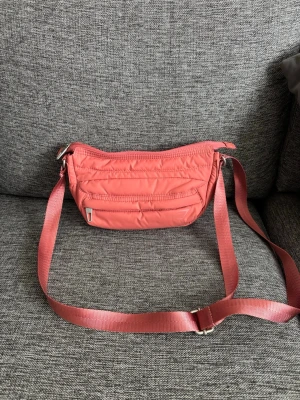 Gymshark axelväska - Snygg rosa axelväska i quiltad design med justerbart axelband. Väskan har en dragkedja upptill och en praktisk ytterficka med dragkedja framtill. Tillverkad i ett lätt och tåligt syntetmaterial. Perfekt för dig som vill ha en färgklick till outfiten.