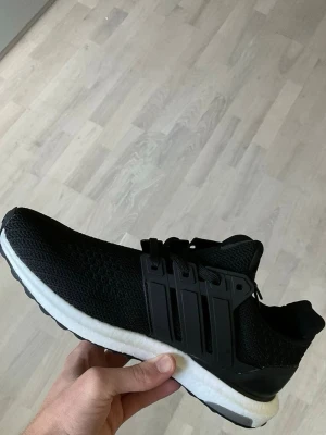 Svarta Adidas Ultraboost sneakers - Säljer ett par svarta Adidas Ultraboost sneakers med stickad ovandel i mesh, klassiska tre ränder på sidan och vit Boost-sula. Skorna har svart plastdetalj vid snörningen och hälkappa för extra stöd. Perfekta för dig som gillar stilrena och bekväma sneakers.