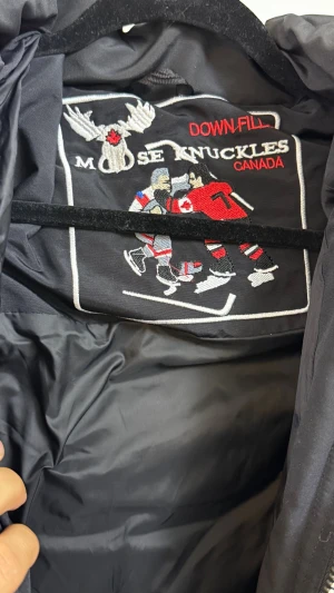 Svart dunjacka från Moose Knuckles - Säljer en svart dunjacka från Moose Knuckles med klassisk logga i metall på ärmen och broderad hockeypatch på insidan. Jackan har en sportig look och är fodrad med dun för extra värme. Perfekt för kalla vinterdagar.