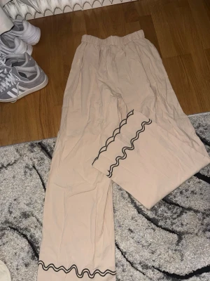 Beiga linnebyxor med vågig kant - Beiga linnebyxor med elastisk midja och raka ben. Byxorna har en svart vågig kantdetalj längst ner på benen som ger en unik och trendig touch. Perfekta för dig som gillar chill och stilren vibe.