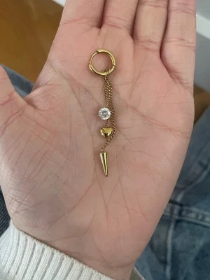 Guldigt hängeörhänge med kedja - Trendiga örhänge i guldton med ring, tunn kedja och tre detaljer: en klar sten, ett litet hjärta och en spetsig kon. 