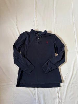 Mörkblå långärmad piké från Polo Ralph Lauren - Klassisk mörkblå långärmad pikétröja från Polo Ralph Lauren med röd broderad logga på bröstet. Tröjan har krage, knappar vid halsen och är gjord i mjuk bomull. Perfekt för en stilren och avslappnad look.