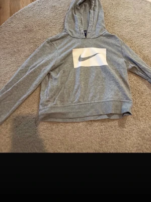 Grå hoodie från Nike med logga - Snygg grå hoodie från Nike med stor vit Nike-logga på bröstet. Hoodien har huva och långa ärmar, tillverkad i mjukt och bekvämt material. Perfekt för en avslappnad och sportig stil.