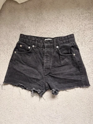 Svarta högmidjade jeansshorts - Säljer ett par svarta högmidjade jeansshorts med råa, fransiga kanter och klassisk femficksdesign. Shortsen har knappar framtill samt bälteshällor. Shortsen är ifrån Pull & Bear och är i storlek EUR 34