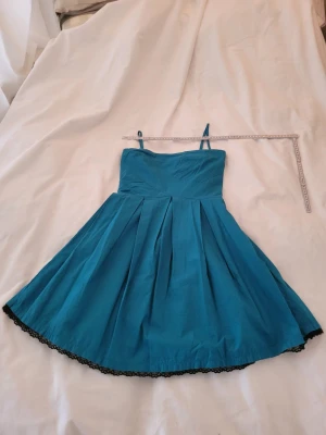 Turkos kort klänning med spetskant - Snygg turkos kort klänning med tunna axelband och utställd kjol. Klänningen har fina veck framtill och en dekorativ svart spetskant längst ner. Perfekt för dig som gillar en feminin och ungdomlig stil.