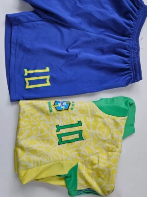 Brasilien fotbollskit nummer 10 - Säljer ett Brasilien fotbollskit med gul tröja och blå shorts. Tröjan har gröna detaljer, nummer 10 och Brasilien-emblem på bröstet. Shortsen är blå med nummer 10 i gult. Perfekt för fotbollsträning eller match.
