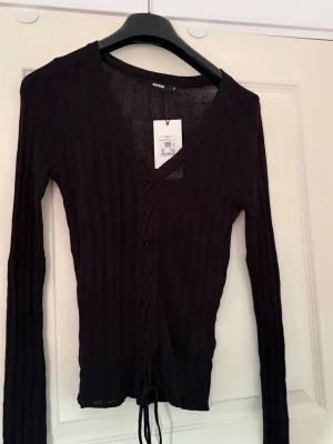 Svart ribbad topp från BikBok - Snygg svart långärmad topp från BikBok med ribbad struktur och snörning framtill. Djup v-ringning och croppad passform som ger en trendig look. Perfekt att styla med höga jeans eller kjol.