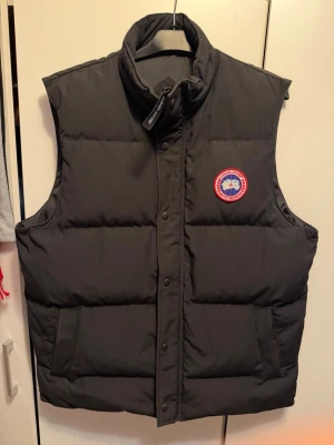 Svart dunväst från Canada Goose - Snygg svart dunväst från Canada Goose med klassisk logotyp på bröstet. Västen har hög krage, tryckknappar och två stora fickor framtill. Perfekt för lager-på-lager och håller dig varm med sitt tjocka dunfoder.