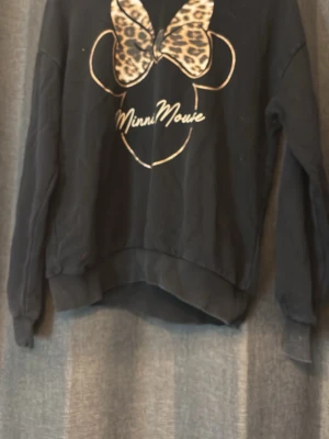 Svart Minnie Mouse hoodie med tryck - Svart hoodie med huva och lång ärm från Disney. Framtill finns ett stort Minnie Mouse-tryck med leopardmönstrad rosett och vit text. Hoodien har ribbade muddar vid ärmslut och nederkant. Perfekt för dig som gillar snygga och lekfulla detaljer.