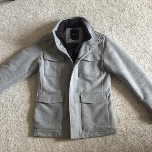 Ljusgrå rock från Jack & Jones - Snygg ljusgrå rock/jacka från Jack & Jones med fyra fickor framtill och hög krage. Dan har en stilren design med dold dragkedja och knappar. Materialet är mjukt och fodret är mörkt för extra komfort. Perfekt för dig som vill ha en klassisk men modern look.