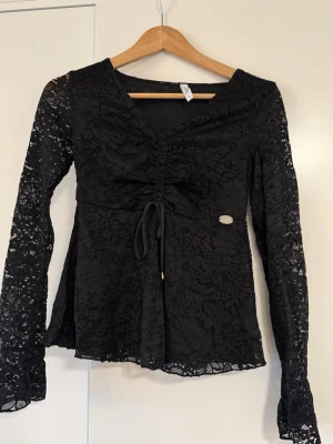 Svart spetsblus med knytning - Snygg svart spetsblus med långa ärmar och v-ringning. Blusen har dekorativ knytning framtill och lätt utsvängd nederdel. Tillverkad i genombruten spets som ger en elegant look. Perfekt att styla med jeans eller kjol.