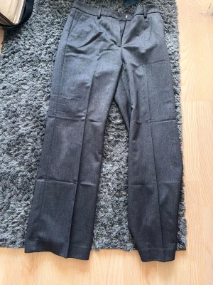 Grå kostymbyxor med pressveck - Snygga grå kostymbyxor med klassiskt pressveck och raka ben. Byxorna har hällor för skärp och stängs med knapp och dragkedja. Perfekta för en stilren och clean look. Materialet känns mjukt och har en lätt glans.