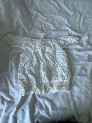 Vit offshoulder topp med spetsdetaljer - Söt vit offshoulder topp med broderade spetsdetaljer längs axlarna och smockad nederkant. Toppen är ärmlös och har en romantisk känsla, perfekt för varma dagar. Passar dig som gillar en feminin och trendig stil.