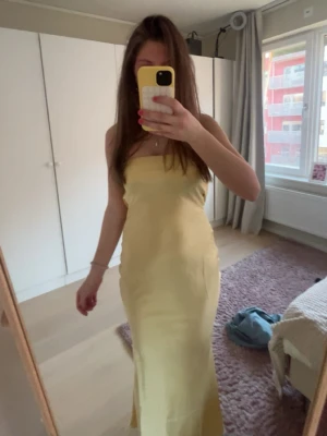 Ljusgul axelbandslös långklänning - Säljer en elegant ljusgul långklänning med axelbandslös design. Klänningen har en smickrande, figurnära passform och är tillverkad i ett mjukt, lätt material som ger ett snyggt fall. Perfekt för sommarens alla tillfällen!