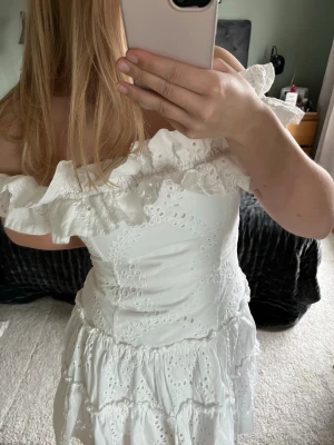 Vit offshoulder klänning med volang - Säljer en söt vit kortklänning i bomull med broderade detaljer och volanger. Klänningen har offshoulder-modell och ärmlös design, samt omlottliknande volang upptill och flera lager i kjolen. Superfin studentklänning. Egentligen m men jag har normalt s