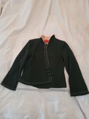Svart omlottjacka med dekorativa knappar - Unik svart omlottjacka med dekorativa knappar och sicksack-broderi längs kanten. Jackan har lång ärm och en snygg, figurnära passform. Insidan har ett kontrasterande foder med mönster. Perfekt för dig som vill sticka ut med detaljer. Köpt i Kina för många år sedan, sparsamt använd. Troligen siden polyester viskos. Väldigt behagligt material.